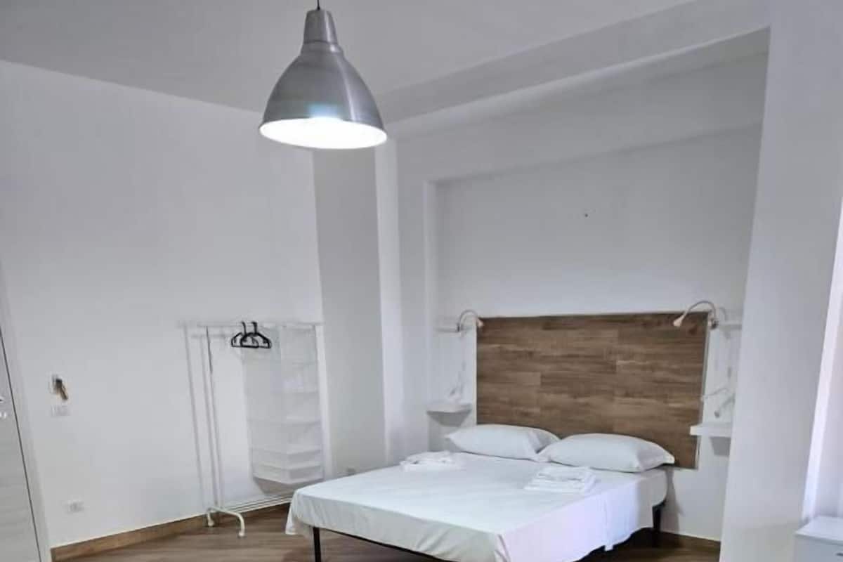A bedroom with a wooden floor, a bed with white bedding, a hanging light fixture, and a white wardrobe.