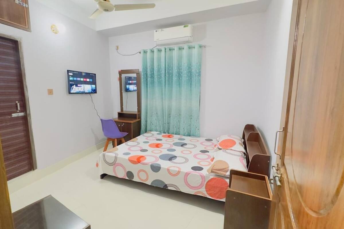 Un dormitorio con cama, televisión, espejo, ventana con cortinas y ventilador de techo.