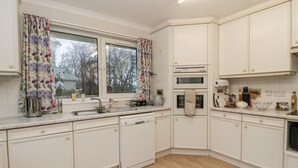 Una cocina con alacenas blancas, lavavajillas, horno y ventana con cortinas florales.