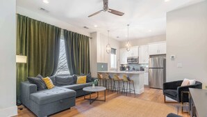A modern living room with a grey sofa, a round coffee table, and a black armchair. A kitchen with white cabinets and stainless steel appliances is visible in the background.