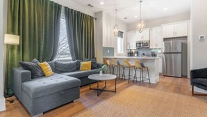 A modern living room with a grey sofa, a small round coffee table, and a kitchen area with white cabinets and stainless steel appliances.