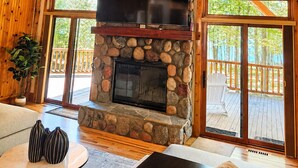 A stone fireplace with a flat-screen TV, wooden beams, and large windows.
