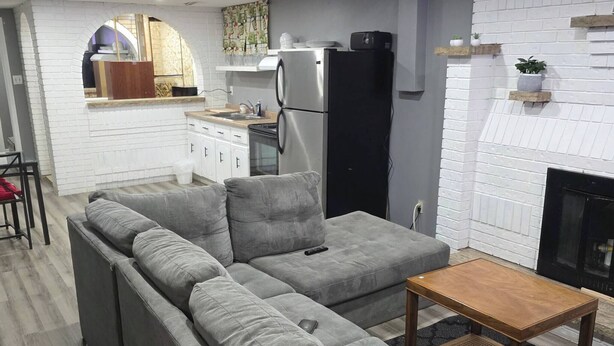 Een moderne woonkamer met een grijze hoekbank, een klein houten bijzettafeltje en een keuken op de achtergrond.