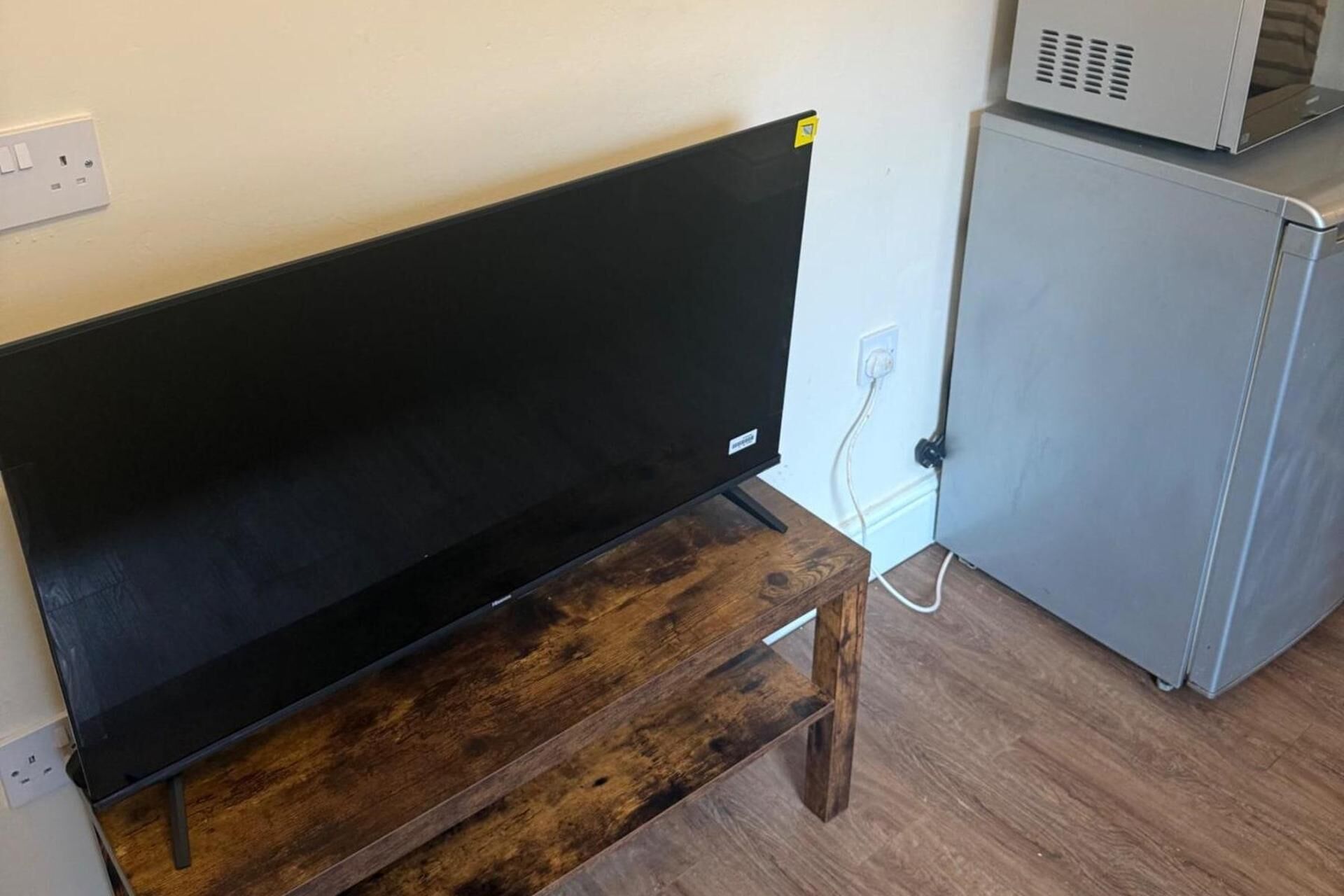 Um quarto com um banco de madeira, uma TV preta em um suporte e uma geladeira branca.
