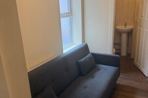Un sofá gris con dos cojines en una habitación con ventana, un lavamanos y una puerta que da al baño.