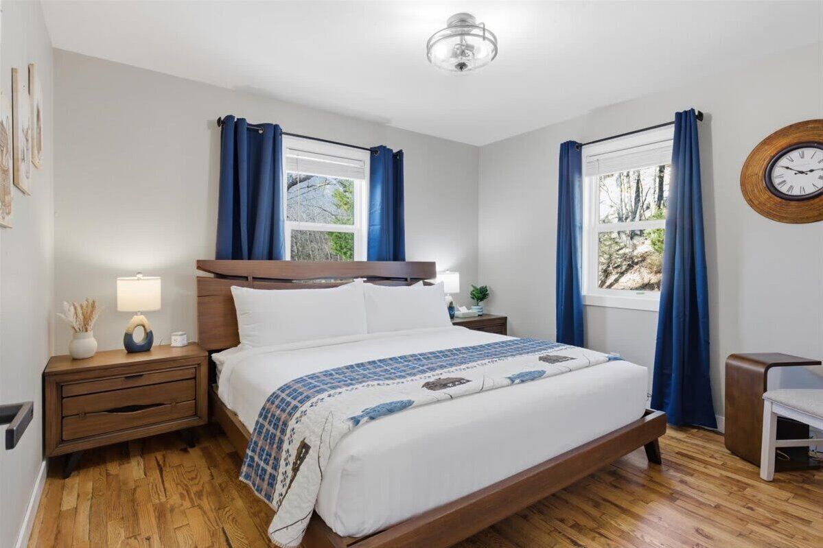 Een slaapkamer met een houten bed, twee nachtkastjes met lampen, een wandklok en een raam met gordijnen.