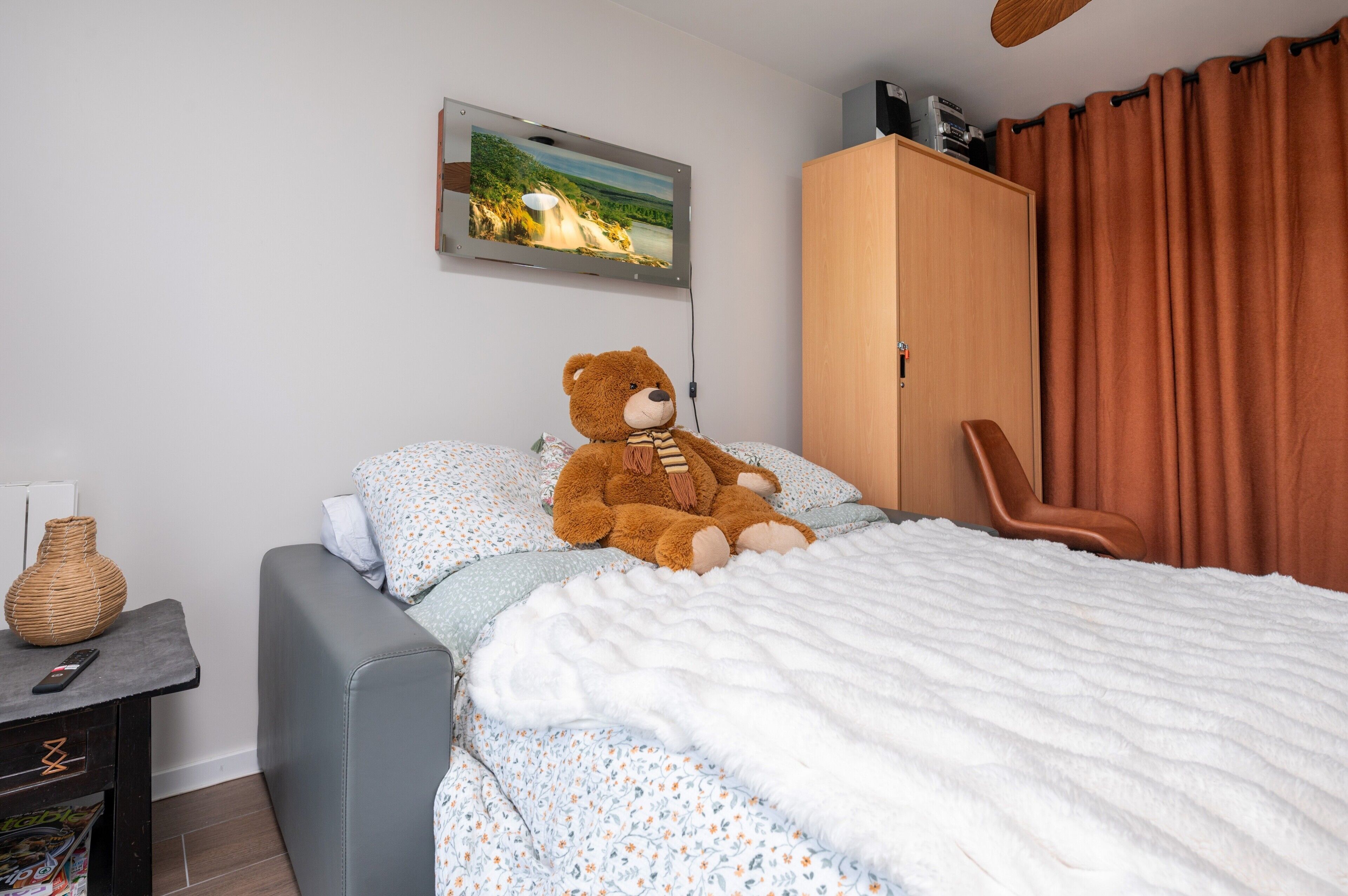 Een bed met een teddybeer, een stoel, een kledingkast, een schilderij en een vaas op een nachtkastje.