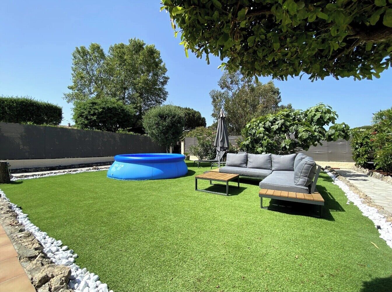 A backyard with artificial grass, a blue inflatable pool, and a grey sofa.
