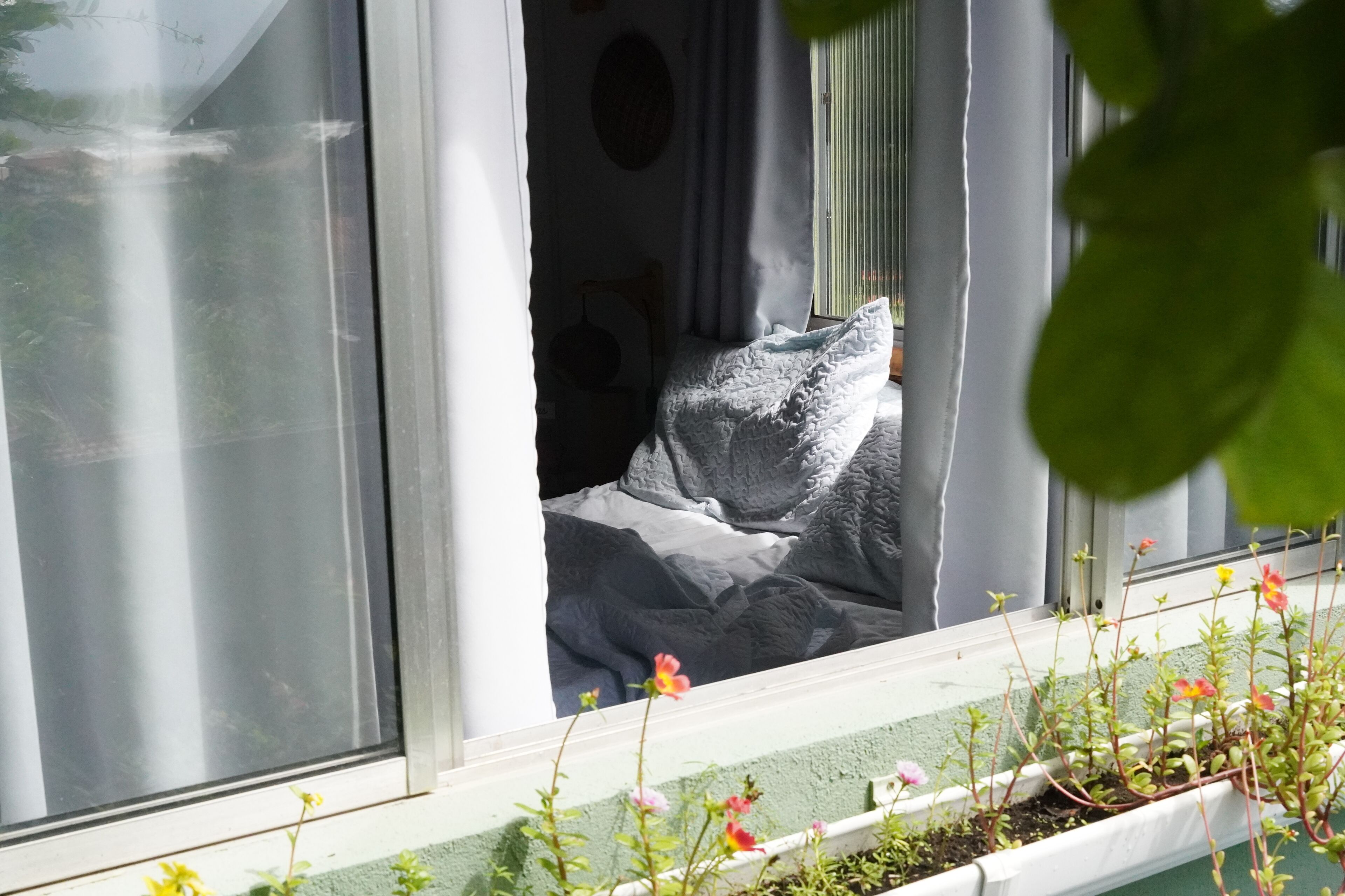 A window with a view of a bed, a curtain, and a plant box with flowers.