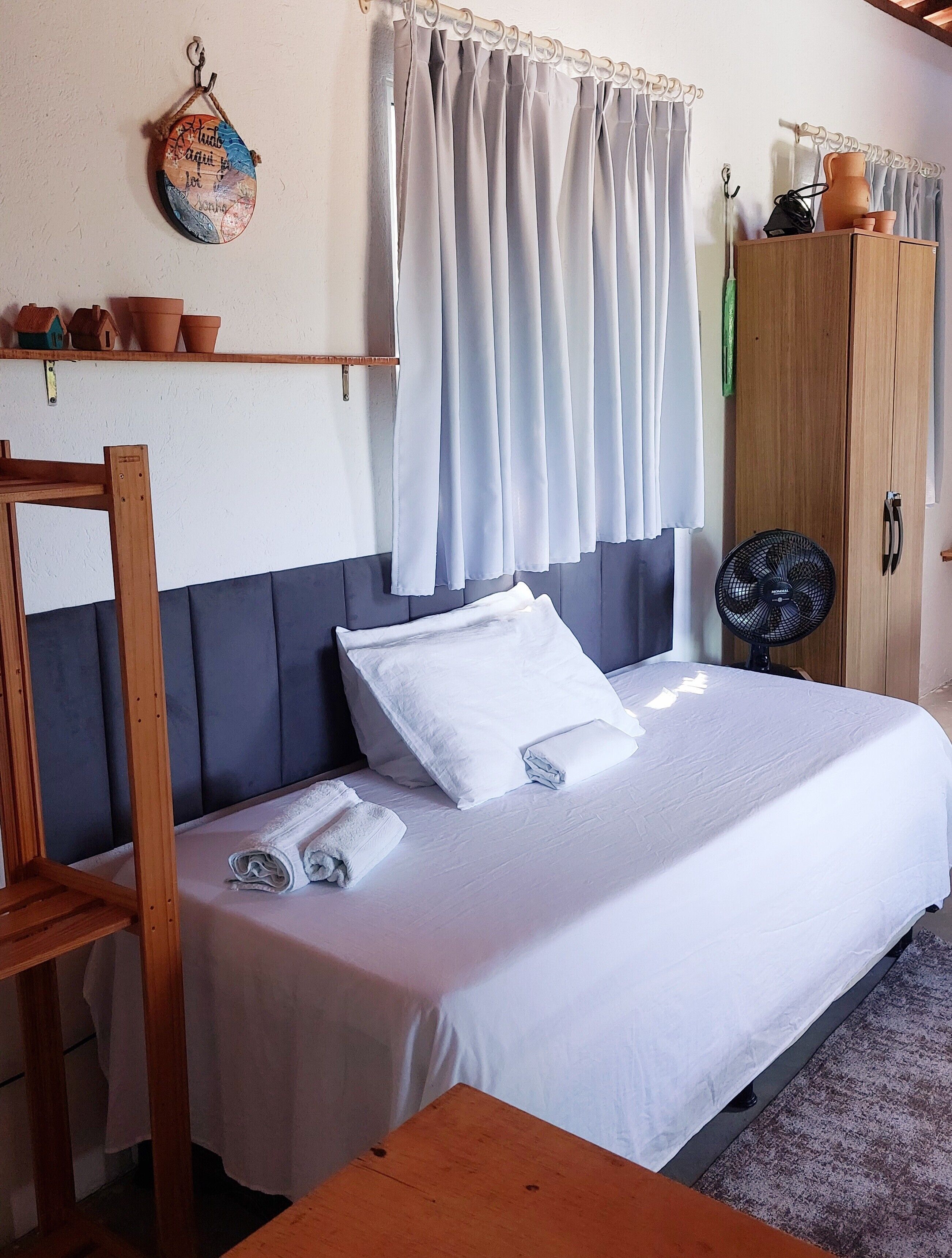 Un lit avec du linge de lit blanc, une tête de lit en bois, une table en bois, un ventilateur et une armoire.