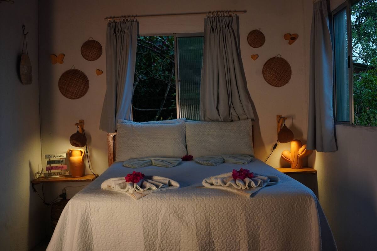 Ein ordentlich bezogenes Bett mit weißer Decke, zwei Kissen und einem dekorativen Kopfteil. Es gibt zwei Nachttische mit Lampen, ein Fenster mit Vorhängen und Wanddekorationen.