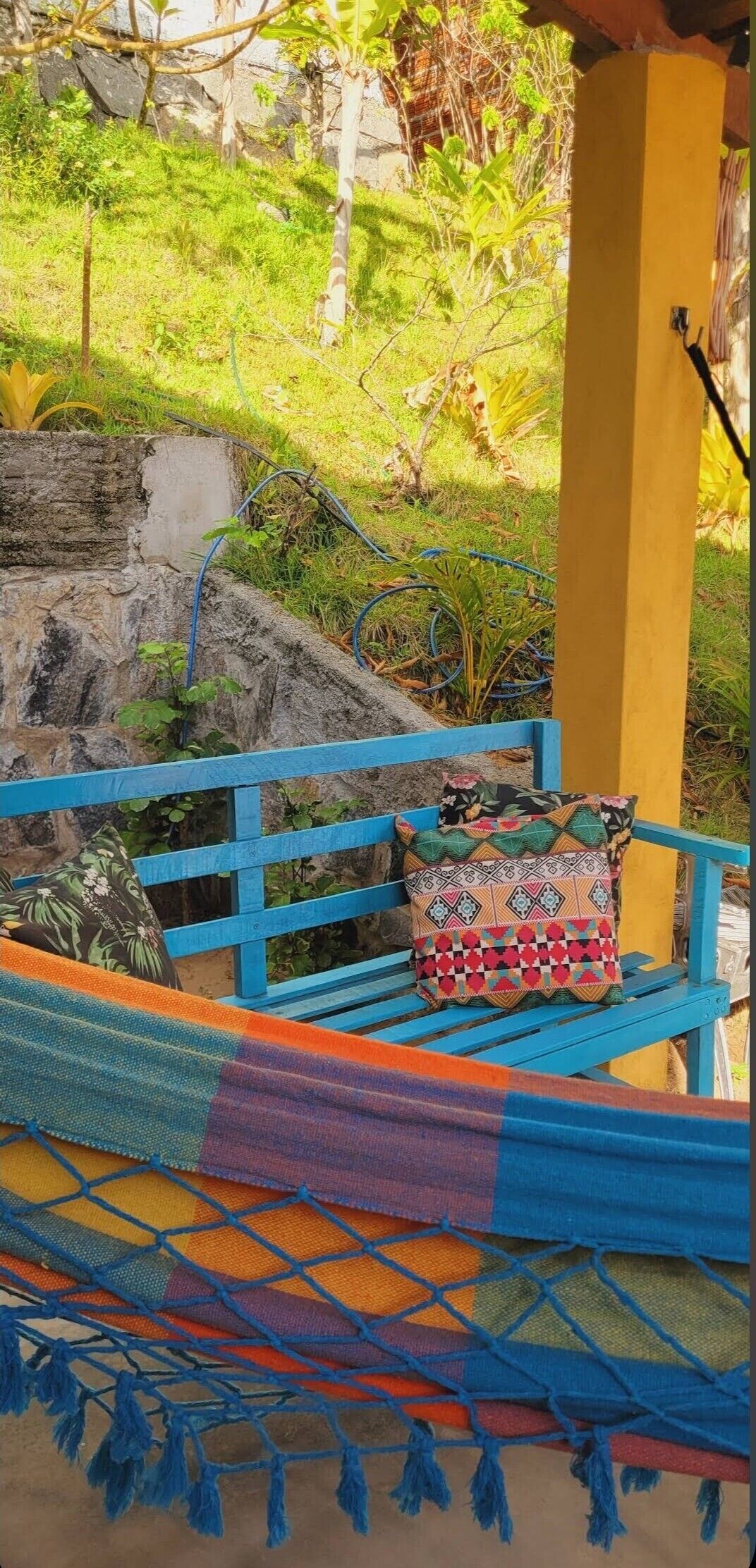 Un hamac fait de tissu coloré et orné de franges, un banc bleu avec un coussin à motifs et un pilier jaune.