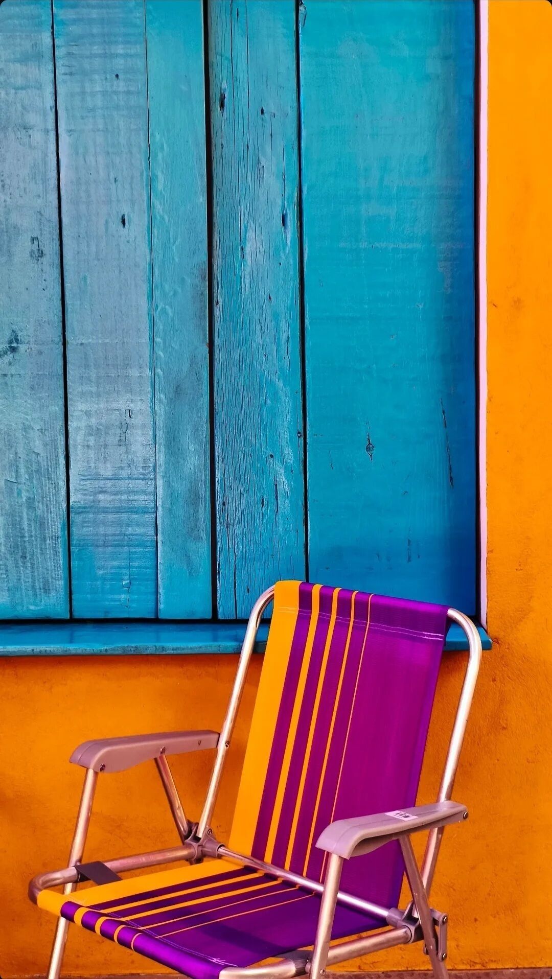 Una silla con respaldo a rayas y estructura metálica, ubicada frente a una ventana con contraventanas de madera azul.