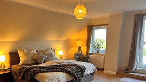 A bedroom with a bed, bedside lamps, a floor lamp, a window with curtains, and a view of the outdoors. - To Huus modern apartment with country view near Verden (Verden (Aller))