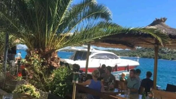 Een boot afgemeerd in de buurt van een strand, met mensen eromheen, palmbomen en een helderblauwe lucht.