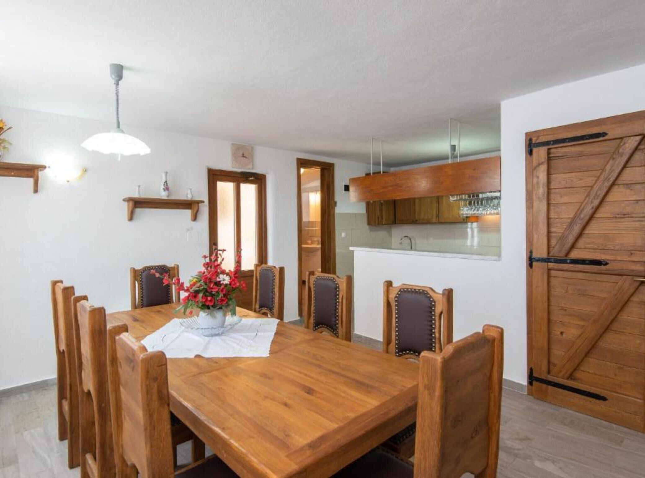 Un comedor con mesa y sillas de madera, un florero, una lámpara colgante y una zona de cocina con armarios de madera y una puerta corrediza.