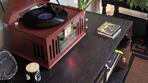 Une platine vinyle en bois, posée sur une table en bois foncé, avec une télécommande, un livre et une petite plante en pot.