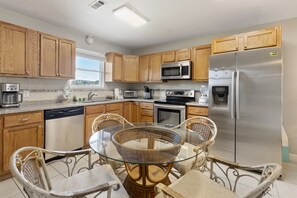 A modern kitchen with wooden cabinets, stainless steel appliances, a glass dining table, wicker chairs, and a view of the outdoors. - Waterfront Secret 2 | Private Beach Access Retreat (Panama City Beach)