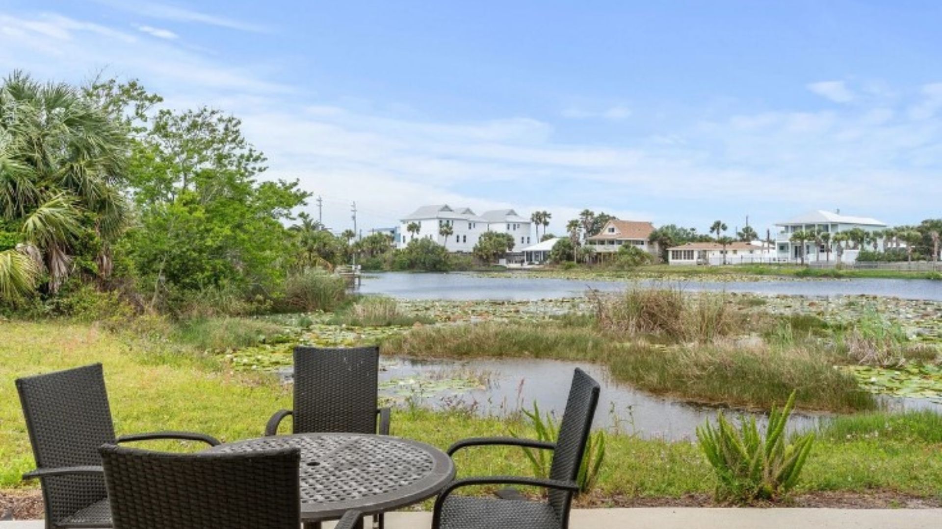 A patio with wicker chairs and a table overlooking a body of water with houses in the background.