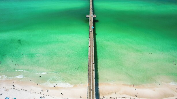 Vista aérea de un muelle que se extiende sobre un mar claro de color verde azulado, con una playa de arena al final.