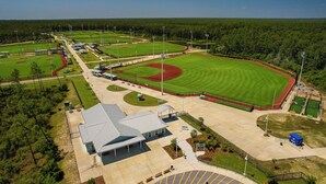 Vue aérienne d’un complexe sportif comprenant plusieurs terrains, un pavillon couvert et un vaste parking.