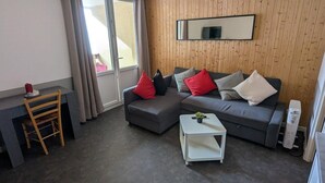 Una sala de estar moderna con un sofá gris, una mesita de noche blanca, una silla de madera y un ventanal con cortinas.