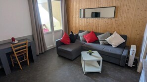 A modern living room with a grey sofa, a small white side table, a wooden chair, and a large window with curtains. - 200m from resort center, 3rd floor, balcony, tv, 27m², Orcières Merlette 1850 (Orcières)