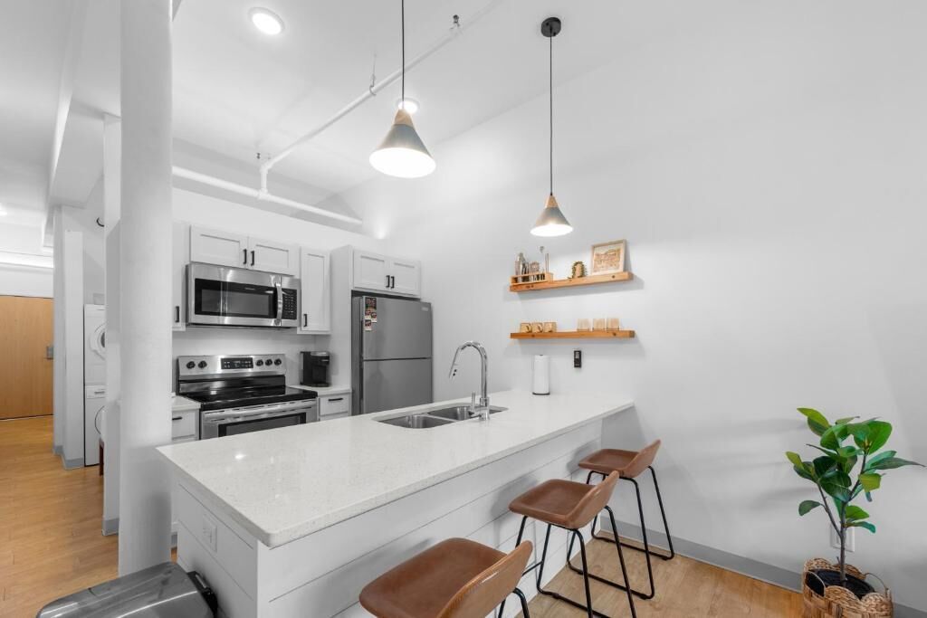 Een moderne keuken met witte kasten, roestvrijstalen apparatuur, een centraal eiland met barstoelen, hangende lampen en een kleine potplant.