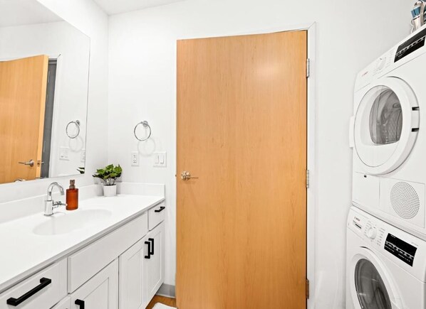 A bathroom with a white sink, wooden door, and stacked washing machines. - Enger Lofts Hotel & Marketplace (Duluth)