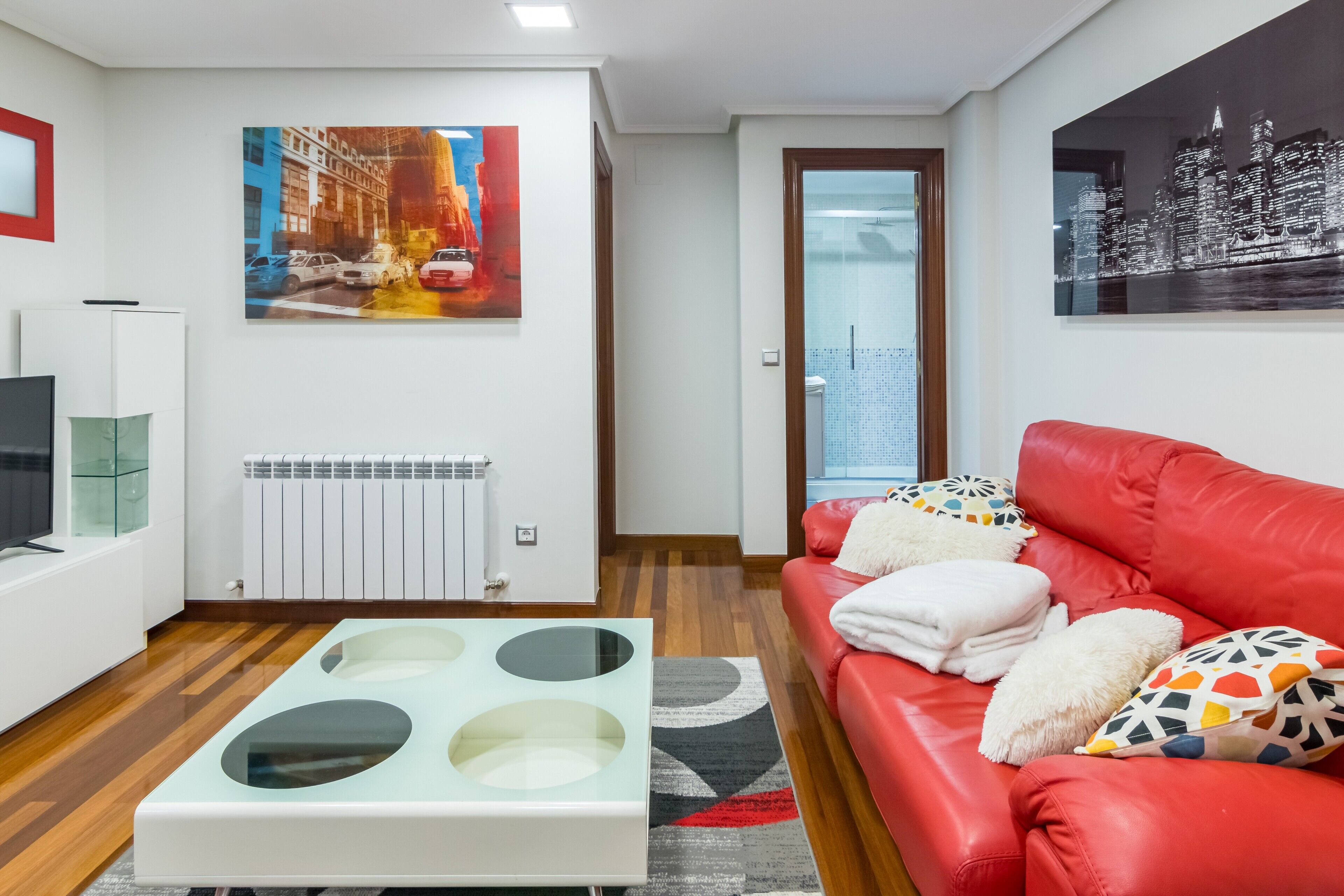 Een moderne woonkamer met een rode leren bank, een glazen salontafel en twee aan de muur gehangen schilderijen.
