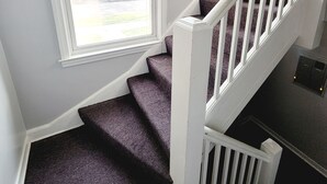 Una escalera con barandilla blanca y alfombra morada, que sube a un rellano con ventana.