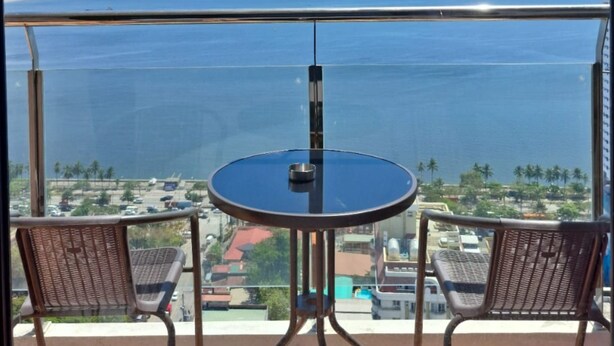 Un balcón con mesa y dos sillas, con vista a un cuerpo de agua donde se ven barcos y una costa lejana.