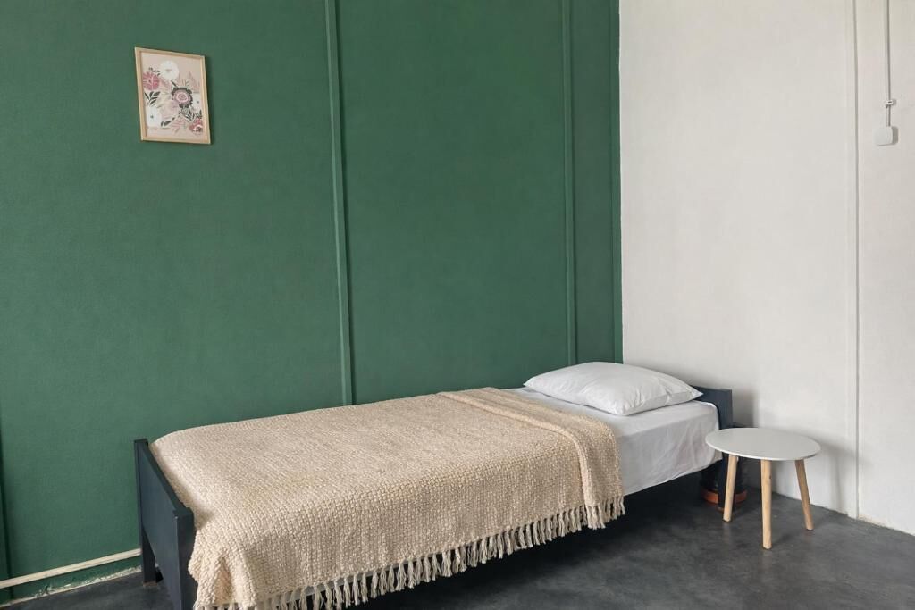 A single bed with a white pillow and a beige blanket, a small round table, and a green wall with a framed picture.