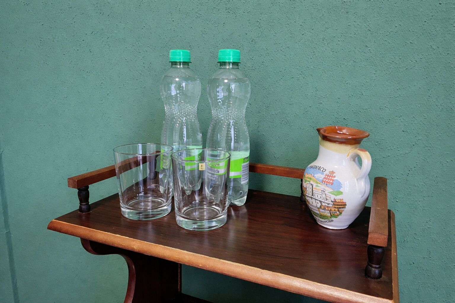 Een houten tafel met drie heldere plastic flessen, twee lege glazen en een keramieken kan.
