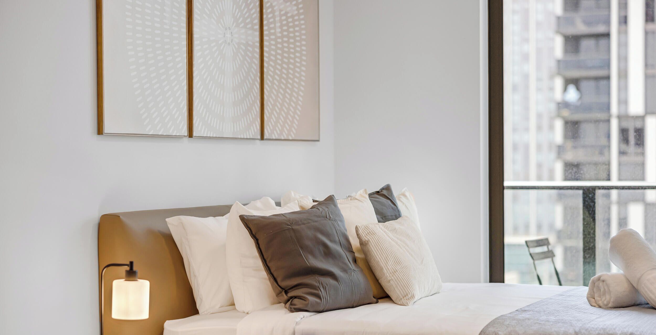 A modern hotel room with a bed, pillows, a wall-mounted light, and a large window with a view of buildings.