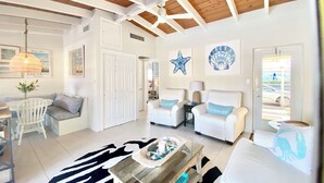 Amplia sala de estar con techo alto, paredes blancas y estructura de vigas de madera. Cuenta con un comedor con mesa y sillas, un sofá con almohadones decorativos y una mesa de centro con una bandeja.
