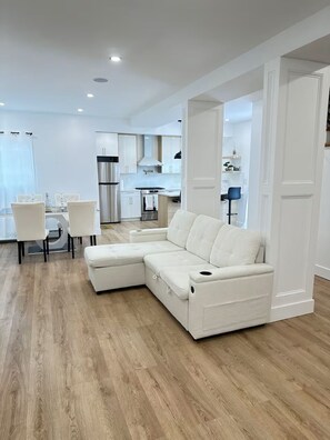 Un salon moderne avec un canapé d’angle, un coin repas avec des chaises et une cuisine en arrière-plan.