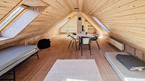 Une petite pièce mansardée, chaleureuse, avec des murs en bois et un plafond en pente. On y trouve un lit, une table avec des chaises et une fenêtre.