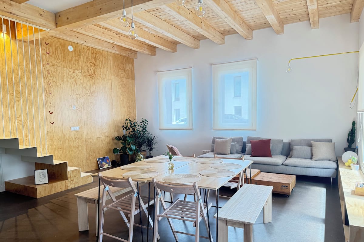 Un interior moderno con una escalera de madera, un comedor con una mesa grande y sillas, un sofá y vistas a través de dos ventanas.