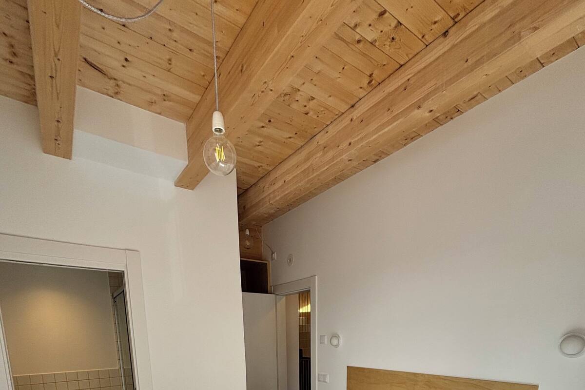 Un dormitorio con techo de madera, una cama con ropa blanca, cabecera de madera y pared blanca.