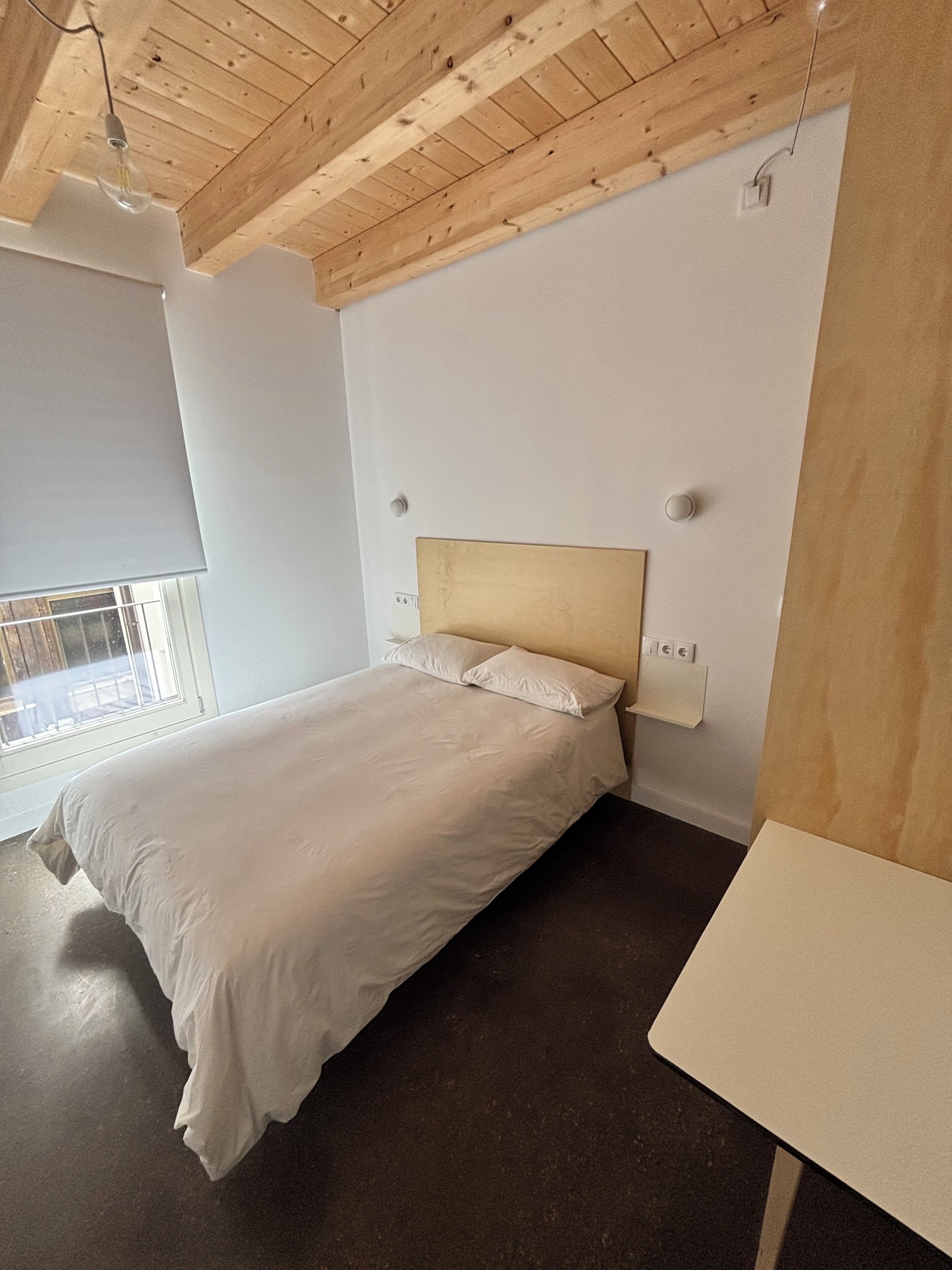Un dormitorio con techo de madera, una cama con ropa de cama blanca, una mesita de noche y una ventana con persianas.