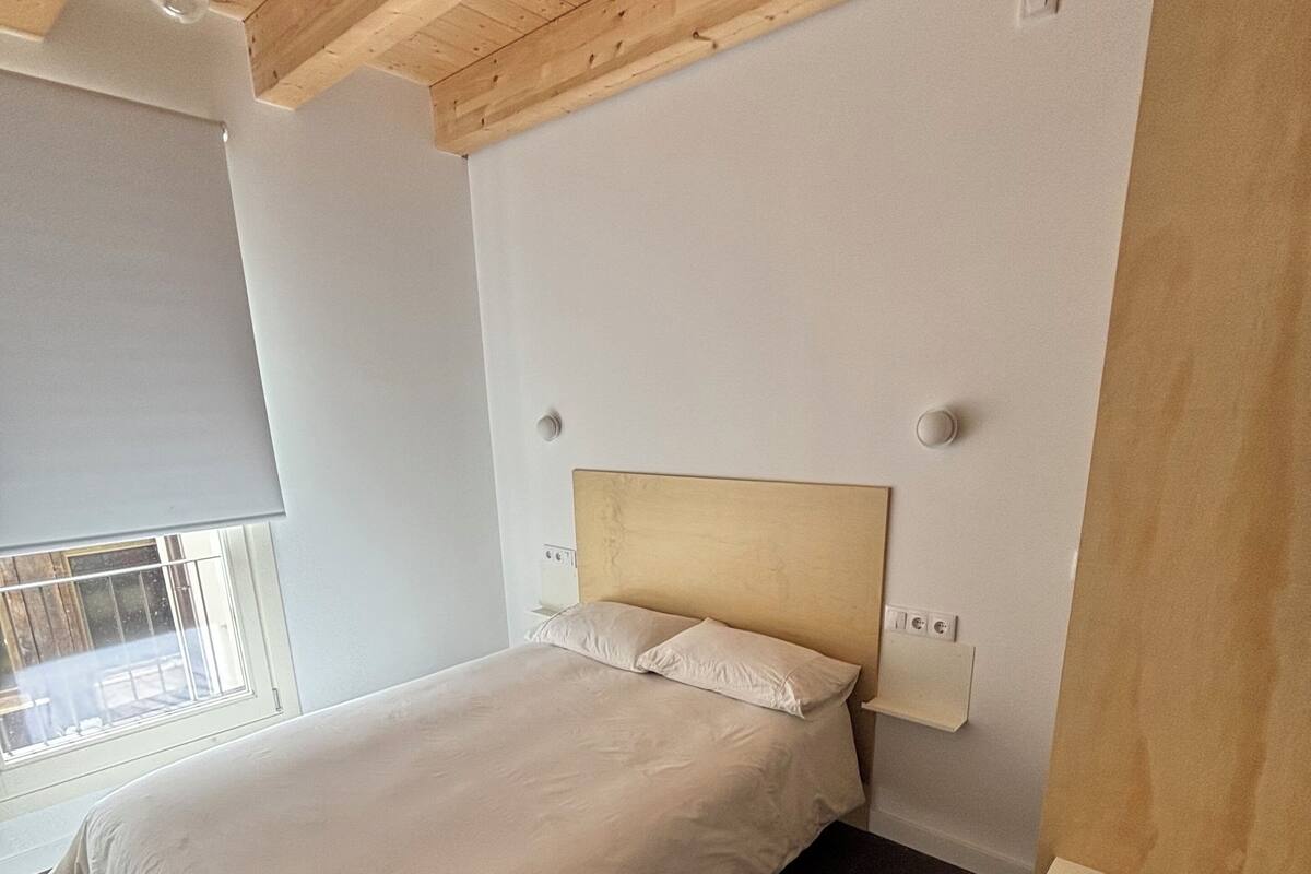 Un dormitorio con techo de madera, una cama con ropa de cama blanca, una mesita de noche y una ventana con persianas.