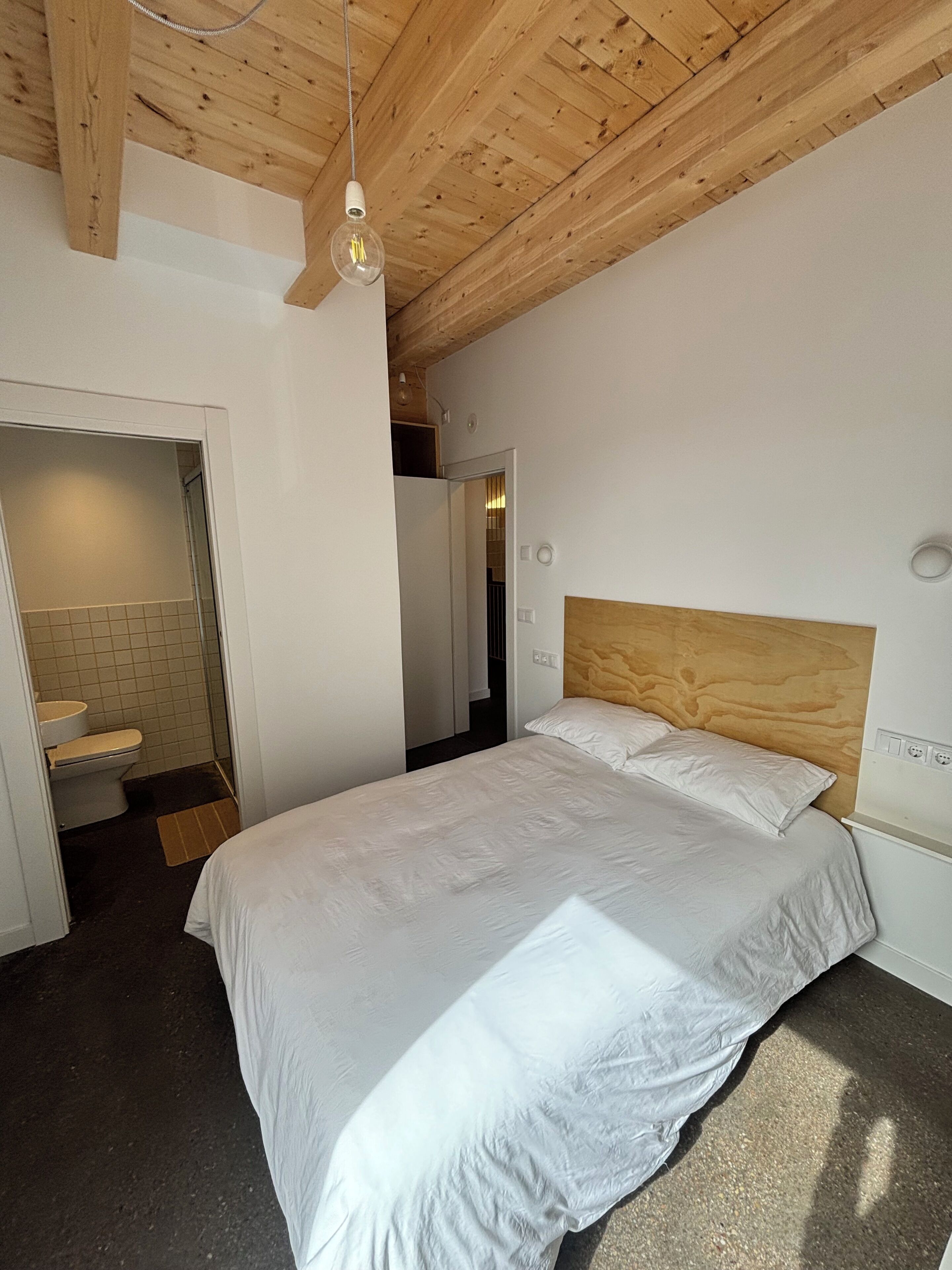 Un dormitorio con techo de madera, cama con ropa de cama blanca, cabecera de madera y pared blanca.