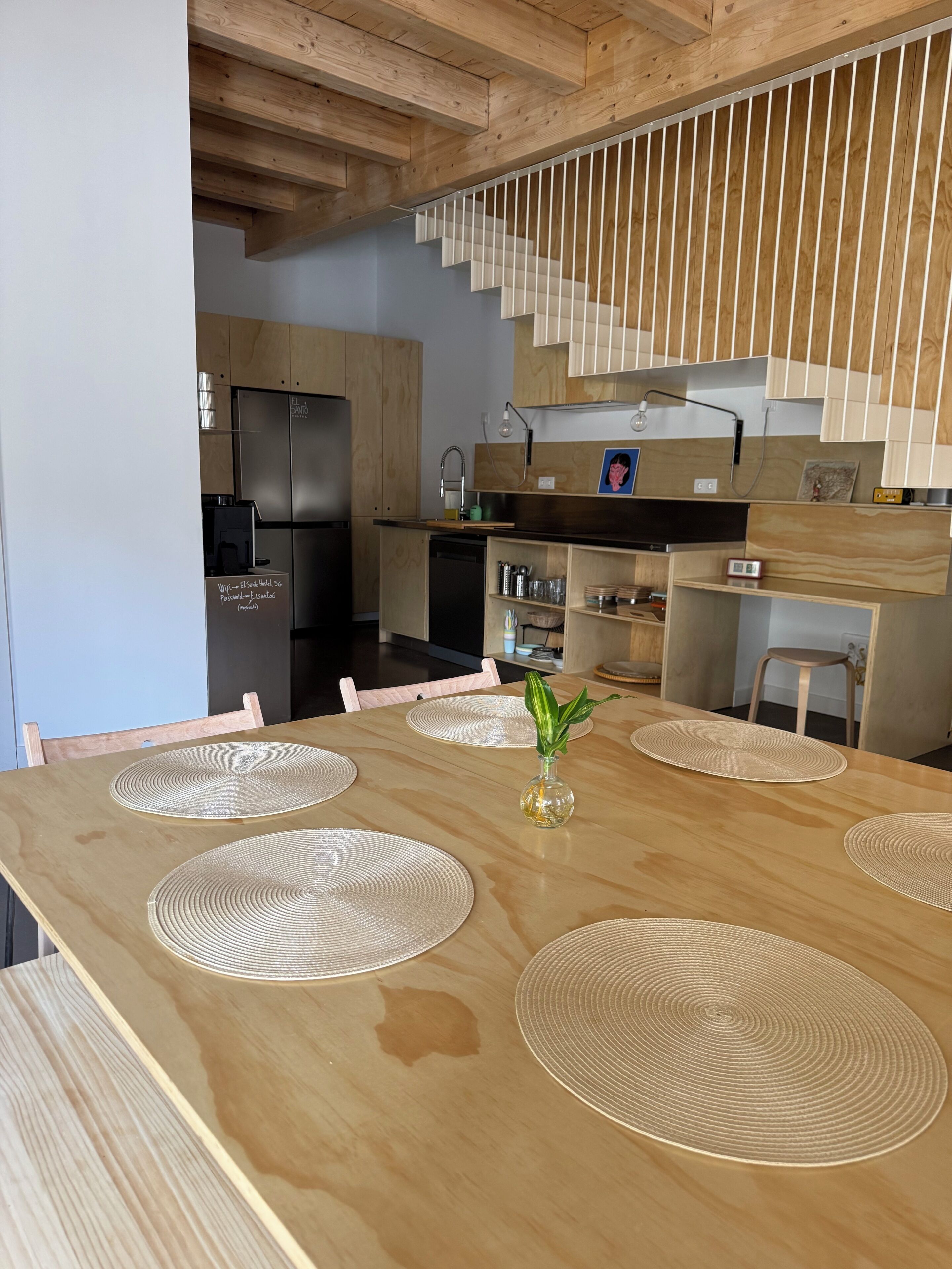 A modern kitchen with wooden cabinets, a stainless steel refrigerator, and a dining area with a wooden table and placemats.