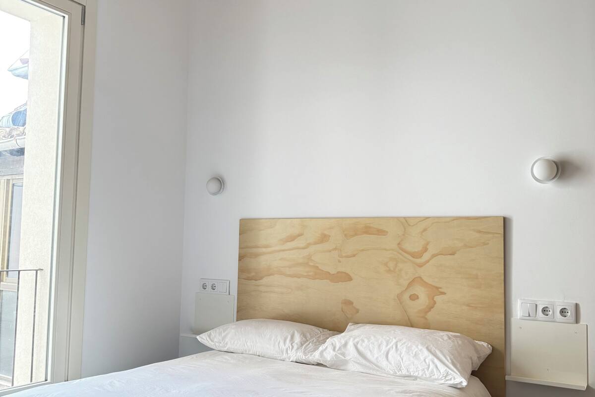 Un dormitorio con cabecera de madera, ropa de cama blanca, una ventana y una lámpara en el techo.