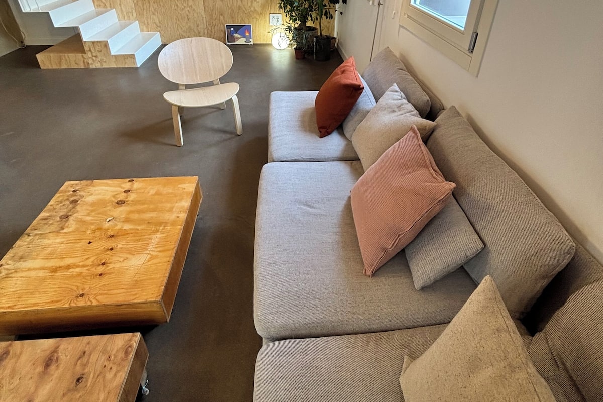 Una sala de estar moderna con una escalera de madera, un sofá gris con cojines, una mesa de centro redonda de madera y una planta en maceta.