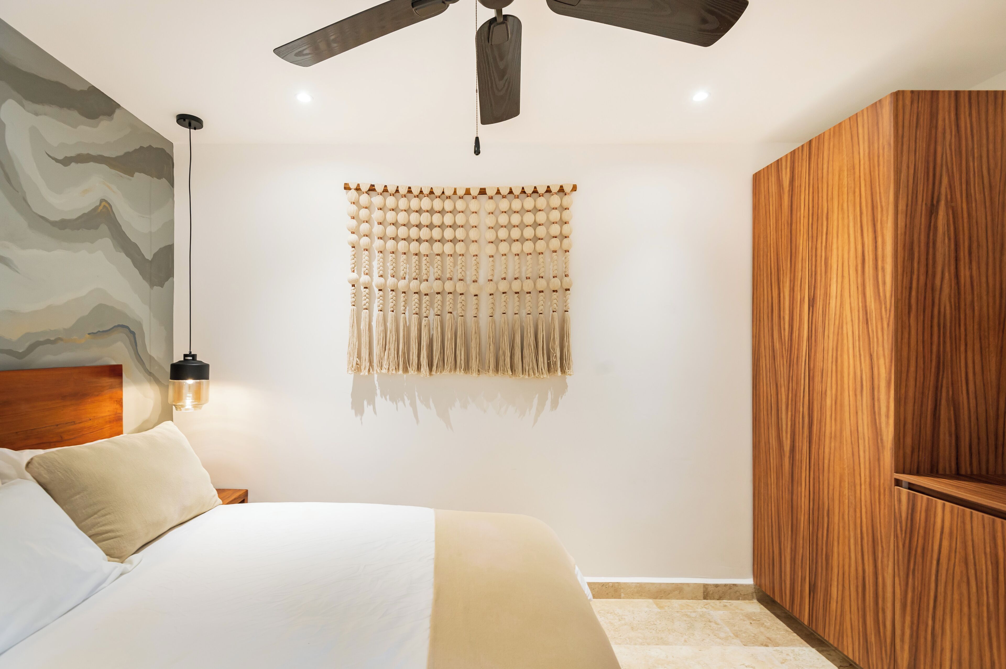 A bedroom with a bed, a ceiling fan, a wall-mounted light fixture, and a wooden wardrobe.