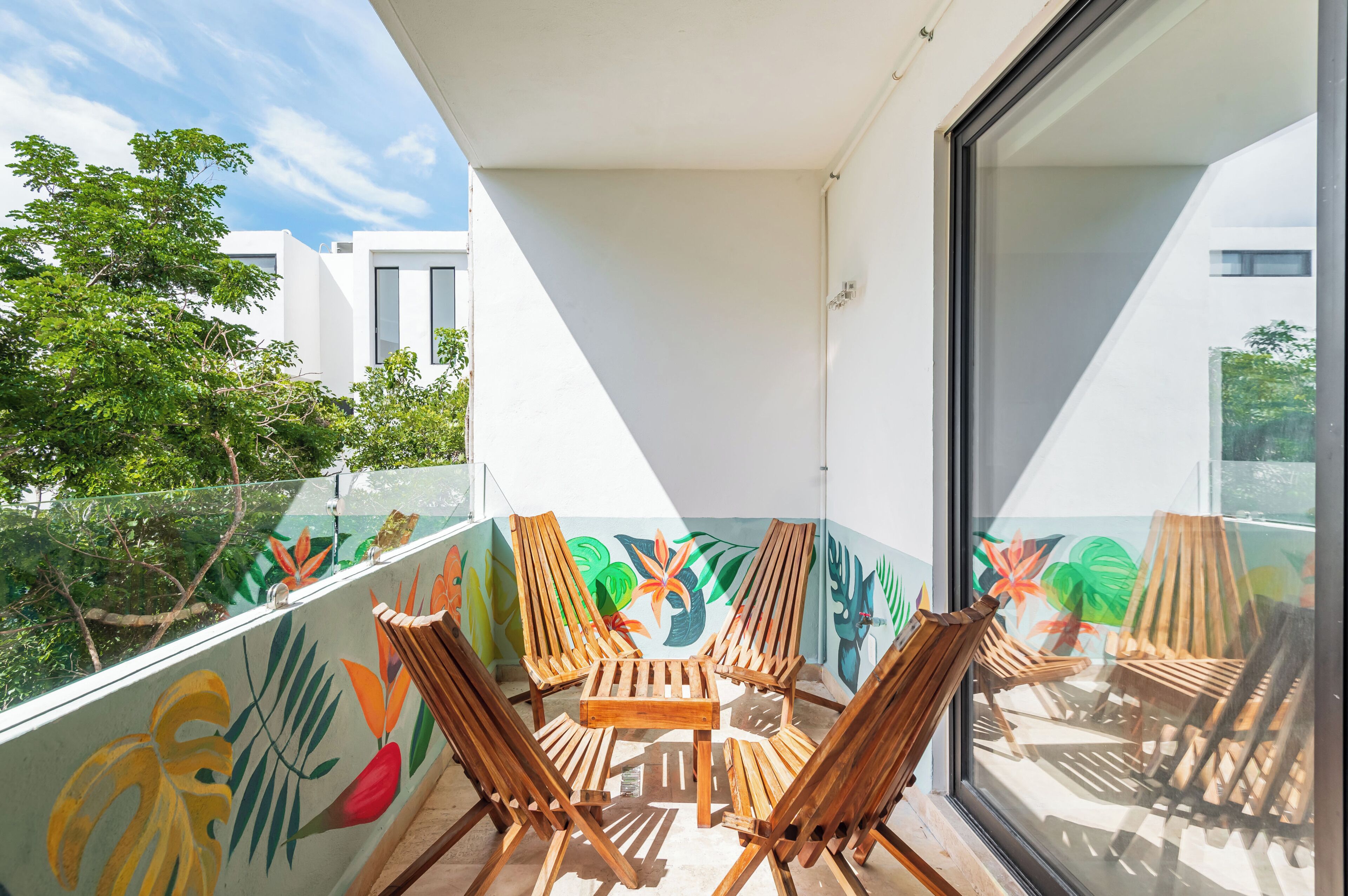 A balcony with wooden chairs and a table, a glass railing, and a mural of tropical leaves.