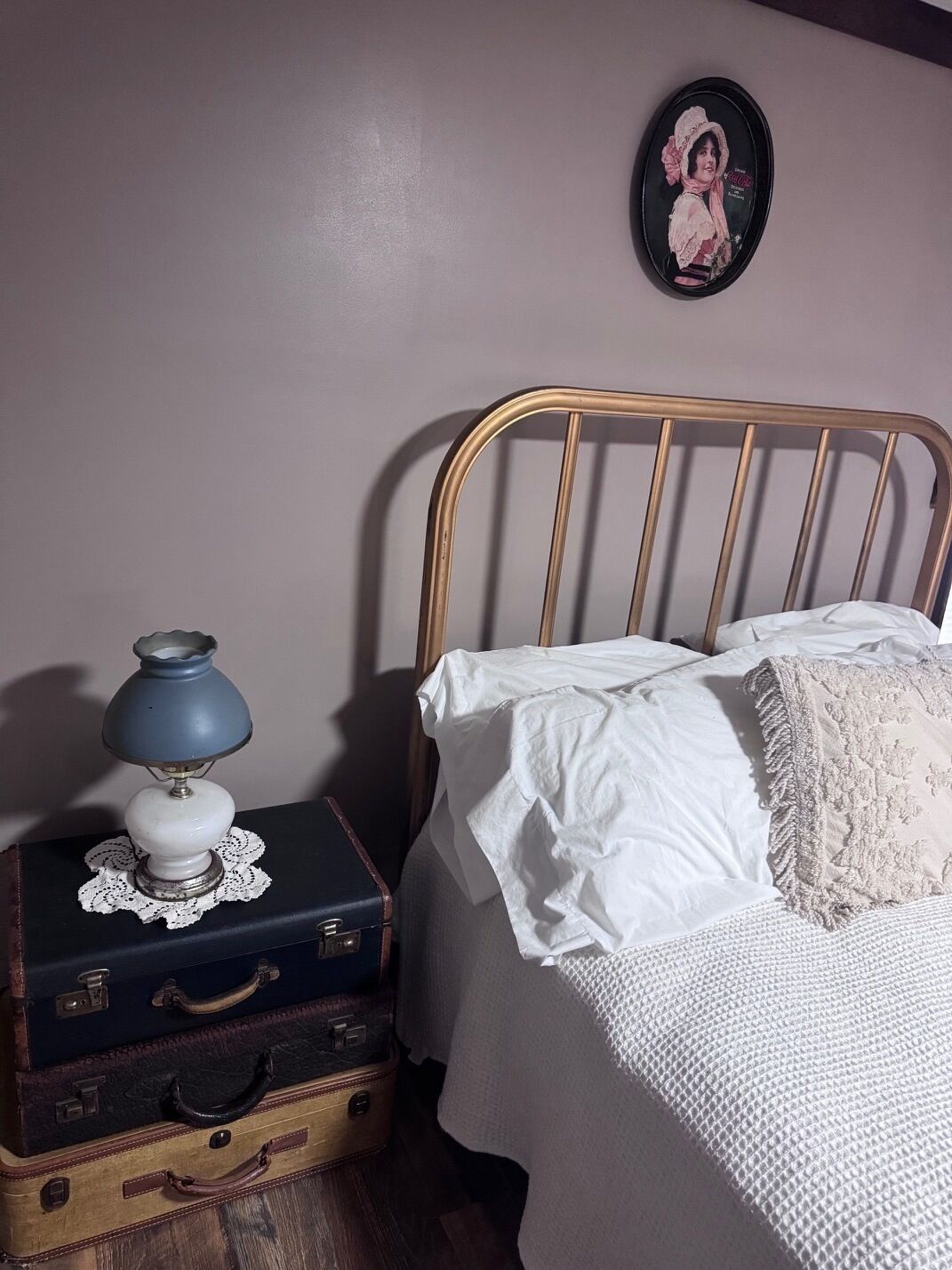 A bed with white linens and a decorative pillow, a vintage lamp on a lace doily atop a suitcase, and a framed picture on the wall.