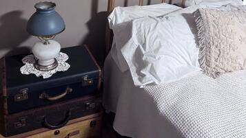 A bed with white linens and a decorative pillow, a vintage lamp on a lace doily atop a suitcase, and a framed picture on the wall.